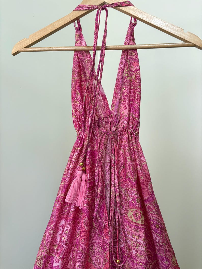 May include: A pink paisley halter dress hanging on a wooden clothes hanger. The dress has a deep V-neck and drawstring waist. A pink tassel hangs from the side. The dress is made of a lightweight fabric.