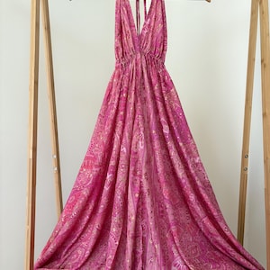 May include: A vibrant pink halter-neck dress with a gold paisley pattern, displayed on a wooden hanger. The dress has a deep V-neck and a flowing, full skirt. The fabric is draped, creating a sense of movement.