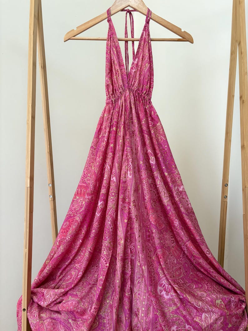 May include: A vibrant pink halter-neck dress with a gold paisley pattern, displayed on a wooden hanger. The dress has a deep V-neck and a flowing, full skirt. The fabric is draped, creating a sense of movement.