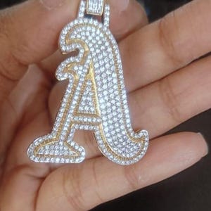 May include: A gold-coloured pendant in the shape of the letter "A", covered in many small, sparkling white gemstones. The pendant is held by a hand, highlighting its detailed design and craftsmanship. The bail is also decorated with gemstones.