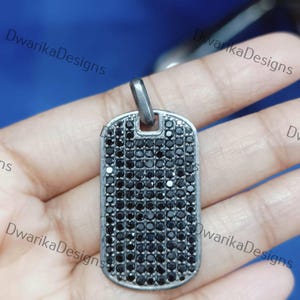 May include: A rectangular pendant with a silver-colored frame and a surface covered in small, black stones. The pendant has a loop at the top for a chain. The design is simple and elegant.