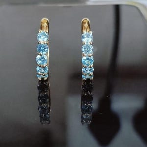 Könnte beinhalten: Ein Paar goldfarbene Creolen, die jeweils mit einer Reihe hellblauer Edelsteine besetzt sind. Die Edelsteine sind gleichmäßig entlang der Krümmung der Creolen angeordnet und erzeugen einen funkelnden Effekt. Die Ohrringe werden vor einem dunklen, reflektierenden Hintergrund gezeigt.