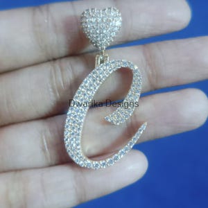 Puede incluir: Un colgante dorado con la letra "C" incrustada con cristales brillantes. Encima de la letra, un diseño en forma de corazón, también cubierto de cristales. El colgante se muestra sobre un fondo azul.