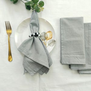 May include: A table setting featuring a gold spoon and fork, a white plate with a floral design, a gray napkin held by a beaded napkin ring, and three folded gray napkins. The setting is on a white tablecloth.