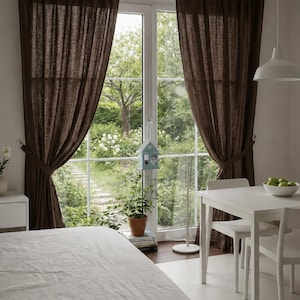 Op de afbeelding: Een lichte kamer met een groot raam omlijst door bruine gordijnen. Een wit bed staat op de voorgrond, en een witte tafel en stoelen staan rechts. Het raam kijkt uit op een weelderige groene tuin.