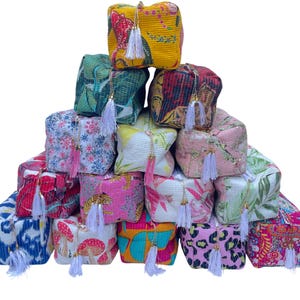 May include: A collection of patterned fabric pouches stacked in a pyramid. Each pouch is square, featuring a tassel and a zip. The designs include floral, geometric, and animal prints in shades of pink, blue, and yellow.