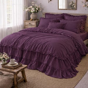 May include: A bedroom with a bed dressed in a deep purple ruffled duvet cover and pillows. A small wooden table holds a tray with a cup and saucer. The room has a neutral colour palette with a large window and a decorative mirror.