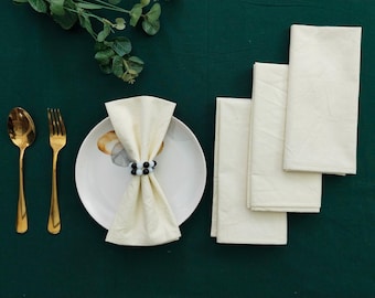Puede incluir: Una mesa puesta con cubiertos dorados, un plato blanco con una servilleta doblada sujeta por un servilletero de cuentas blancas y negras, y varias servilletas crema dobladas. El conjunto está sobre una superficie verde oscuro.