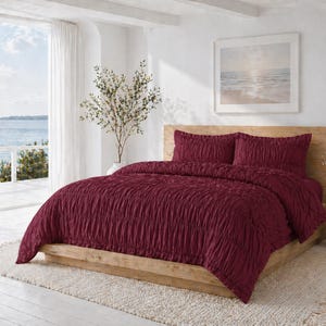 May include: A burgundy bedspread and two matching pillowcases on a wooden bed frame. The bedding has a textured, ruched design. The room has a coastal theme with a view of the ocean through a large window.