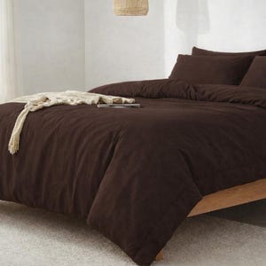 Può includere: Un letto con un copripiumino marrone scuro e federe abbinate. Una coperta color crema e un libro sono appoggiati sul letto. Il telaio del letto è in legno chiaro.