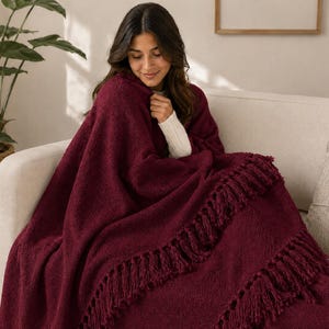 May include: A deep burgundy knitted throw blanket featuring a textured surface and long, decorative fringe detailing along the edges. The blanket is draped over the shoulders and lap, showcasing the thick, soft yarn construction and rich colour.