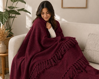 May include: A deep burgundy knitted throw blanket featuring a textured surface and long, decorative fringe detailing along the edges. The blanket is draped over the shoulders and lap, showcasing the thick, soft yarn construction and rich colour.