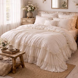 May include: A cream-colored bed with ruffled bedding, pillows, and a matching duvet cover. The bedroom features a wooden nightstand, a small wooden table, and a jute rug. A vase of flowers sits on the nightstand.