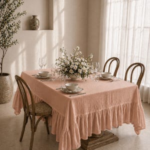 Op de afbeelding: Een eettafel gedekt met een blush roze tafelkleed met ruches. De tafel is gedekt met witte borden, kommen en glaswerk. Rotan stoelen omringen de tafel en een bloemstuk voegt een decoratief tintje toe.