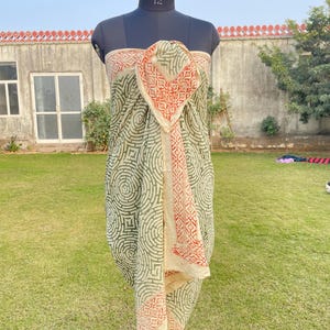 May include: A beige and olive green sarong with a geometric pattern and a red and beige border. The sarong is draped on a mannequin. The background is a grassy lawn and a building.