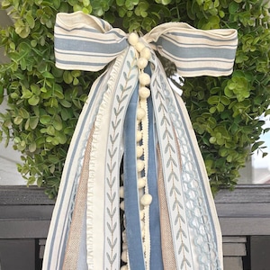 May include: A decorative wreath with a green leafy base and a large bow made of striped fabric in shades of blue and cream. The wreath is adorned with multiple ribbons in various textures and colors, including blue velvet and cream lace.