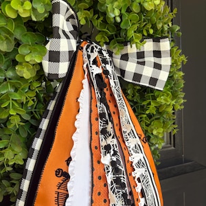 May include: A Halloween-themed wreath with a black and white checkered bow and orange and black ribbons. The ribbons feature spiderwebs, ghosts, and a haunted house design. The wreath is set against a backdrop of green foliage.