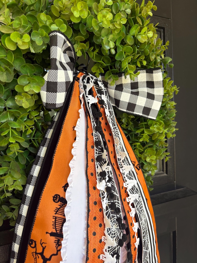 K&ouml;nnte beinhalten: Ein Halloween-Wreath mit einer schwarz-wei&szlig; karierten Schleife und orangefarbenen und schwarzen B&auml;ndern. Die B&auml;nder zeigen Spinnweben, Geister und ein Spukhaus-Design. Der Kranz befindet sich vor einem Hintergrund aus gr&uuml;nem Laub.