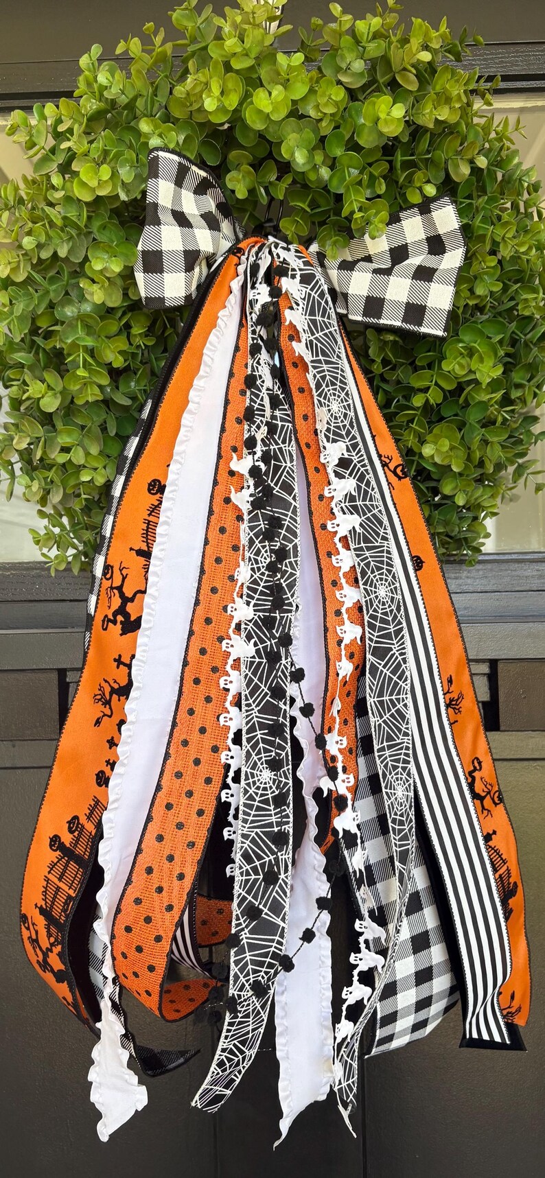 K&ouml;nnte beinhalten: Ein Halloween-Kranz mit gr&uuml;ner Basis und einer schwarz-wei&szlig; karierten Schleife. Der Kranz hat orangefarbene und schwarze B&auml;nder mit Halloween-Mustern, darunter Flederm&auml;use, Spinnweben und Tupfen. Wei&szlig;e B&auml;nder mit Spitzenbesatz sorgen f&uuml;r Kontrast.