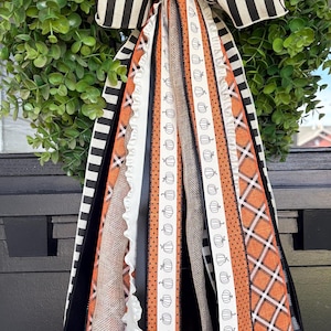 May include: A decorative bow featuring long, cascading ribbons in various textures and patterns, including black and white stripes, solid black, natural burlap, white with small orange pumpkin prints, and orange with a white plaid pattern.