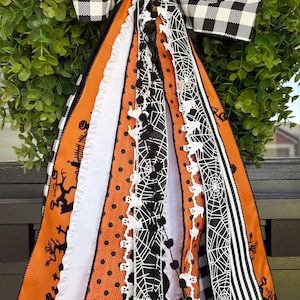 K&ouml;nnte beinhalten: Ein Halloween-Kranz mit gr&uuml;ner Basis und einer gro&szlig;en schwarz-wei&szlig; karierten Schleife. Der Kranz hat orange, schwarze und wei&szlig;e B&auml;nder mit Halloween-Mustern, darunter Geister, Spinnweben und Tupfen.