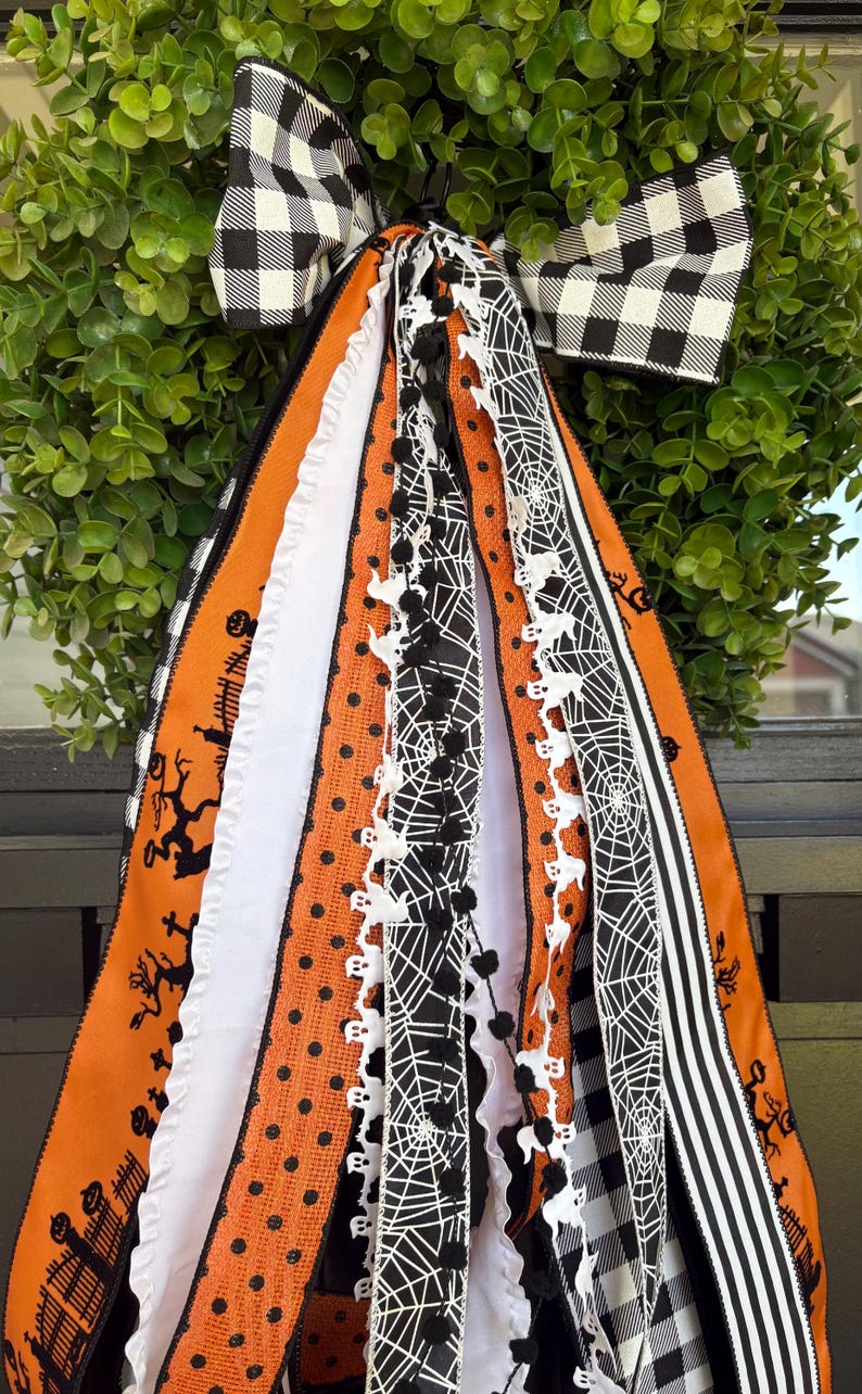 K&ouml;nnte beinhalten: Ein Halloween-Kranz mit gr&uuml;ner Basis und einer gro&szlig;en schwarz-wei&szlig; karierten Schleife. Der Kranz hat orange, schwarze und wei&szlig;e B&auml;nder mit Halloween-Mustern, darunter Geister, Spinnweben und Tupfen.