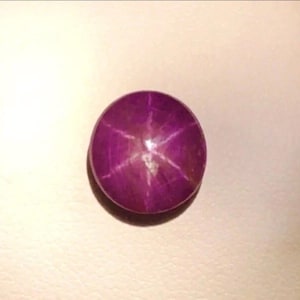 May include: A round, cabochon-cut star ruby gemstone with a deep magenta-purple hue. The stone features a distinct six-rayed star pattern, known as asterism, which radiates from the centre point across the smooth, polished surface of the gem.