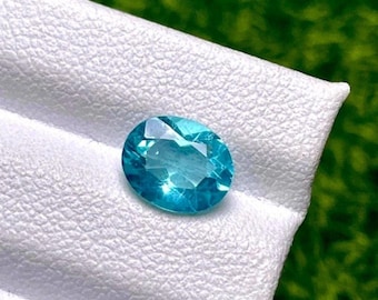 May include: A faceted, oval-shaped, teal gemstone. The gem is set against a white, textured surface, with a blurred green background. The gemstone's facets reflect light, creating a sparkling effect. The gem is a vibrant shade of teal.