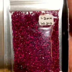 Pode incluir: Um saco de plástico transparente cheio de pequenas e brilhantes pedras preciosas vermelho escuro. Uma pequena etiqueta branca no saco diz "1,2 mm" e "103,21 ct". O saco está dentro de um recipiente prateado.