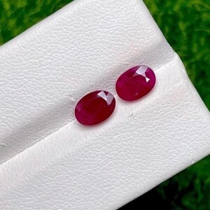 May include: Two oval-cut, deep red rubies with faceted surfaces. The gemstones are positioned side-by-side on a white, textured display surface. Each ruby exhibits a translucent quality with light reflecting off the geometric facets.
