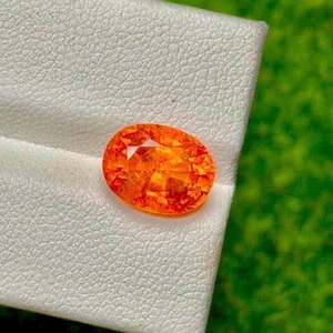 May include: An oval-shaped, bright orange gemstone with a faceted surface. The gem is set against a white textured background, with a blurred green backdrop. The gemstone's colour ranges from bright orange to a deeper reddish-orange.