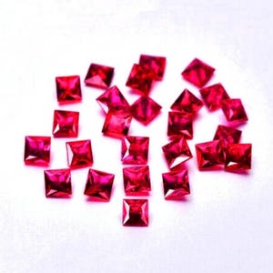 May include: A collection of small, square-cut, vibrant red gemstones. Each stone features a faceted surface that reflects light, creating a brilliant appearance. The stones are scattered across a plain, light-colored surface in a loose arrangement.