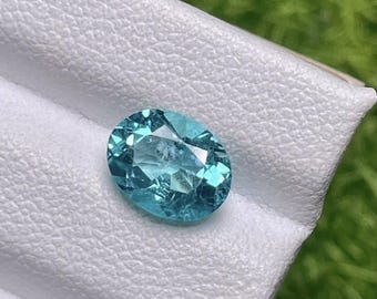 May include: An oval-cut, light blue gemstone with facets, resting on a white, textured surface. The gemstone reflects light, showcasing its clarity and color. The background is a blurred green, suggesting a natural setting. This gemstone is suitable for jewelry.
