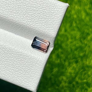 Pode incluir: Uma pedra preciosa retangular com um gradiente de cores, incluindo azul, pêssego e castanho. A gema está colocada sobre uma superfície branca texturizada, com um fundo verde. A gema é provavelmente uma turmalina ou uma pedra preciosa semelhante.