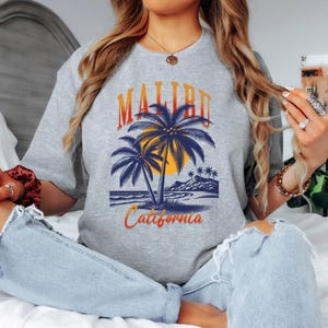 Könnte beinhalten: Ein graues T-Shirt mit einem Grafikdesign von Palmen, einem Sonnenuntergang und den Worten "Malibu California" in Orange und Blau. Das T-Shirt ist ein lässiger Stil, geeignet für den Alltag.