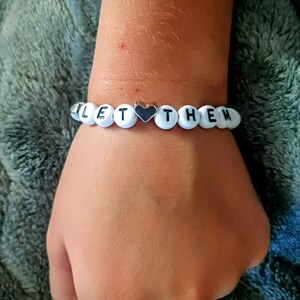 May include: A white beaded bracelet spelling "LET THEM" with black letters and a silver heart charm. The bracelet is worn on a wrist against a soft, textured background.