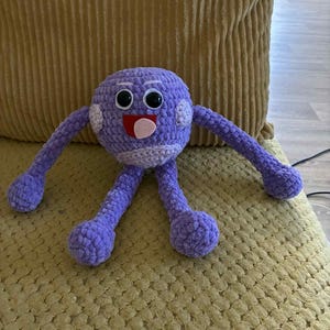 May include: A handmade purple crochet monster toy with googly eyes, a red mouth, and white accents. The plush toy has long arms and legs, and is sitting on a yellow cushion. This whimsical creature is perfect for children.