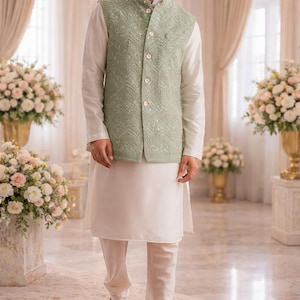 May include: A man's formal wear ensemble. The outfit includes a light green embroidered vest over a white long-sleeved shirt and matching white pants. The look is completed with brown embroidered loafers. The background features floral arrangements.