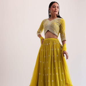 以下が含まれることがあります： 黄緑色のツーピースのフォーマルな衣装。トップスはVネックで、3/4袖に白い刺繍が施されています。スカートは長く流れるようなデザインで、繊細な白い花の模様とフリルの裾が付いています。お揃いのスカーフが腕にかけられています。