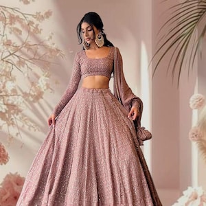 Puede incluir: Conjunto lehenga choli bordado en color rosa empolvado. El conjunto incluye un top corto de manga larga y una falda fluida, ambos con intrincados diseños. El atuendo se complementa con grandes pendientes dorados. El logotipo "IMPERIOEARTH" es visible.