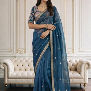 May include: A teal saree with gold embroidered trim and scattered gold accents. The blouse features intricate floral embroidery. The saree is draped over a woman, showcasing the elegant design and flowing fabric.
