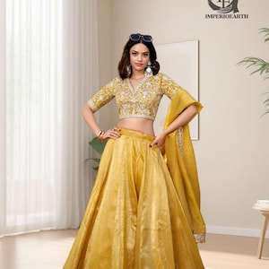 Op de afbeelding: Mosterdgeel formeel ensemble met een crop top en een zwierige rok. De top heeft ingewikkelde borduursels en korte mouwen. Een bijpassende sjaal is over de arm gedrapeerd. Een zonnebril ligt op het hoofd. De outfit is geschikt voor speciale gelegenheden.