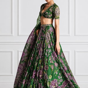 May include: A green and purple floral embroidered lehenga choli set. The set features a V-neck blouse with short sleeves and a flowing skirt. The outfit is adorned with intricate embroidery and sequins. The model is wearing the outfit.