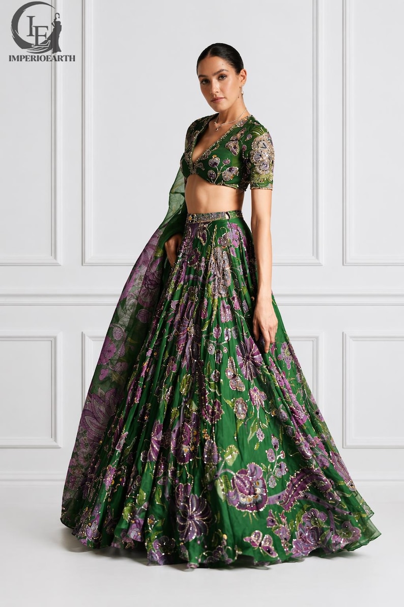May include: A green and purple floral embroidered lehenga choli set. The set features a V-neck blouse with short sleeves and a flowing skirt. The outfit is adorned with intricate embroidery and sequins. The model is wearing the outfit.