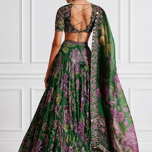 May include: A green floral print lehenga with a matching blouse and dupatta. The lehenga features a full skirt with purple and gold accents. The blouse has a lace-up back. The dupatta has a floral pattern and a beaded border.