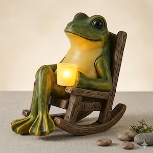 Peut inclure: Une figurine de grenouille verte et jaune assise dans une chaise à bascule brune texturée. La grenouille a de grands yeux noirs et tient dans ses mains une petite lumière cylindrique jaune qui émet une lueur douce et chaleureuse.