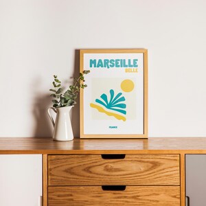 May include: A framed art print with the words "MARSEILLE BELLE FRANCE" in teal and yellow. The print features a stylized beach scene with a yellow sun, teal water, and yellow sand. A white pitcher with greenery sits to the left.