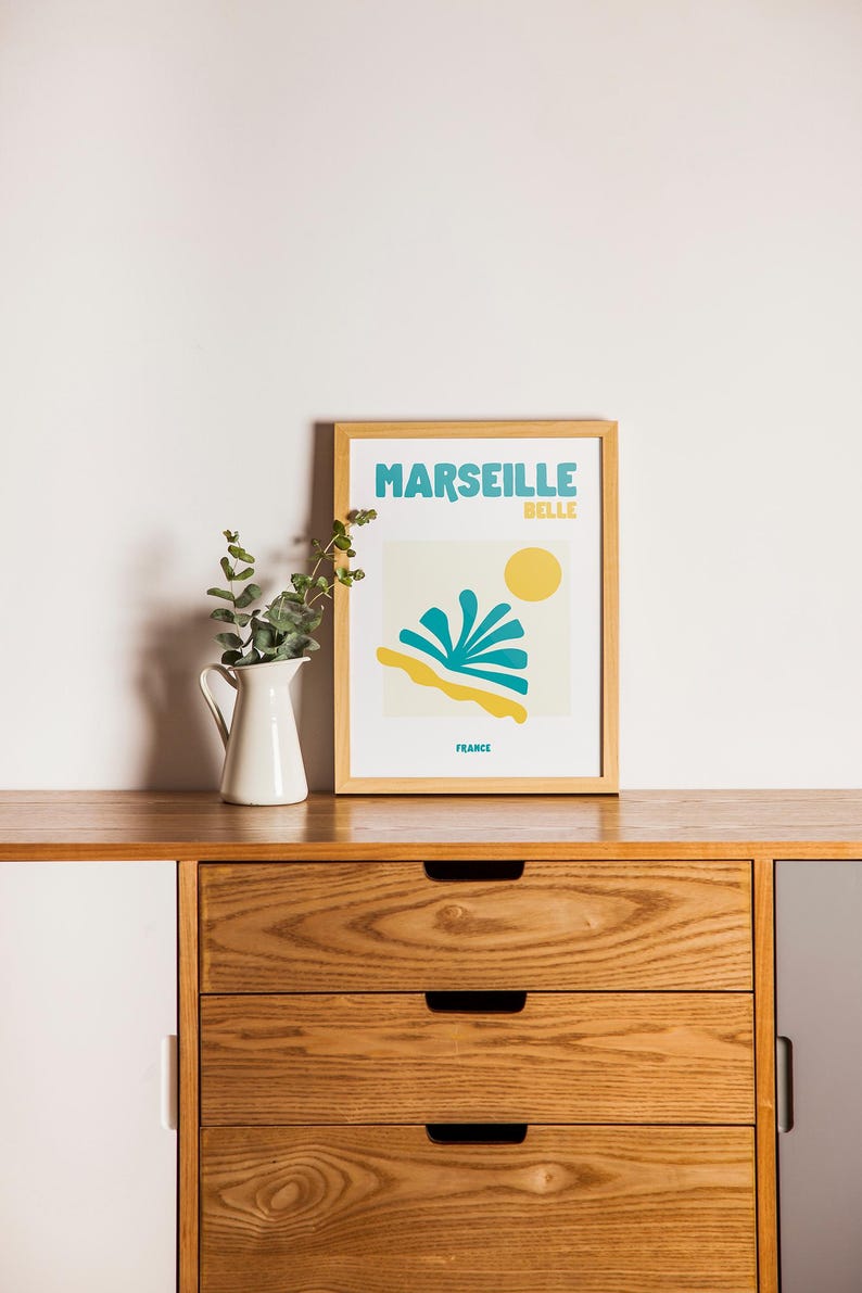May include: A framed art print with the words "MARSEILLE BELLE FRANCE" in teal and yellow. The print features a stylized beach scene with a yellow sun, teal water, and yellow sand. A white pitcher with greenery sits to the left.