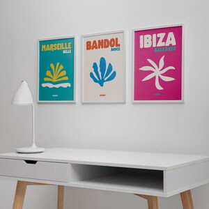 May include: Three framed travel art prints on a white wall. The prints feature colorful abstract designs and the names "Marseille", "Bandol", and "Ibiza". A white desk with a lamp is in the foreground.