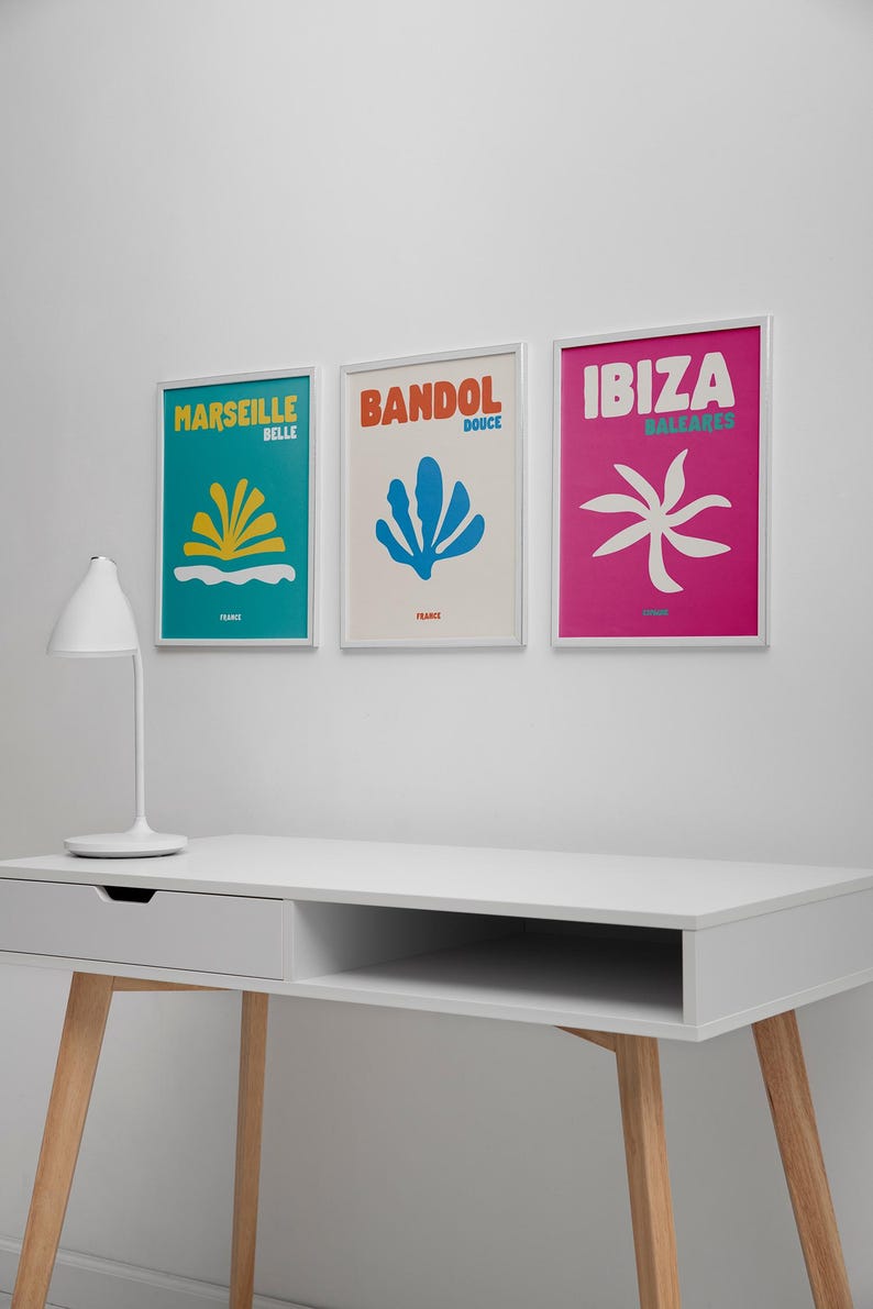 May include: Three framed travel art prints on a white wall. The prints feature colorful abstract designs and the names "Marseille", "Bandol", and "Ibiza". A white desk with a lamp is in the foreground.